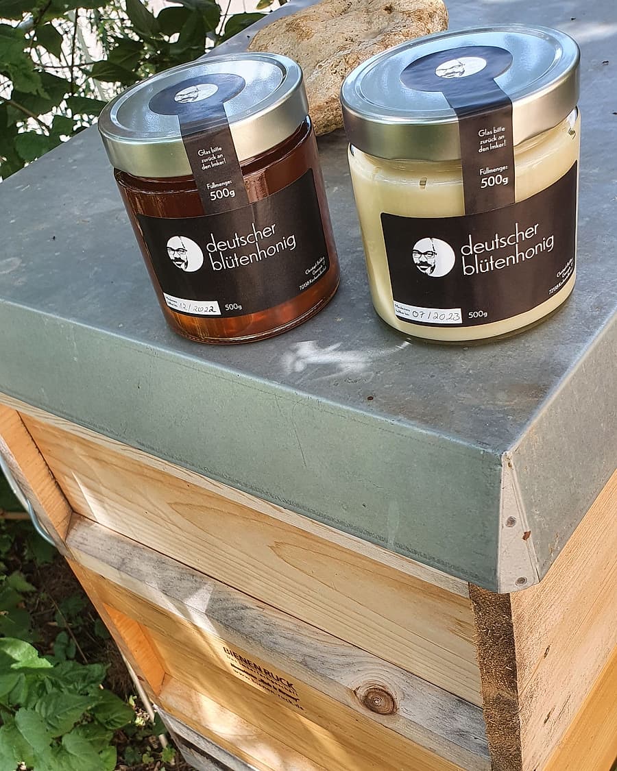 Zwei 500-Gramm-Gläser Blütenhonig, eines dunkel, eines hell und cremig, stehen auf dem Blechdeckel einer Bienenbeute im Garten.